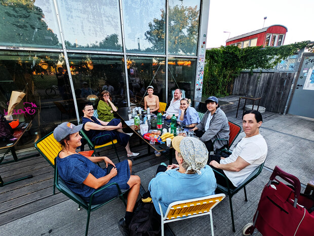 10 Personen sitzen rund um einen Tisch auf einer Terasse (des Flucc) und schauen großteils in RIchtung Kamera.